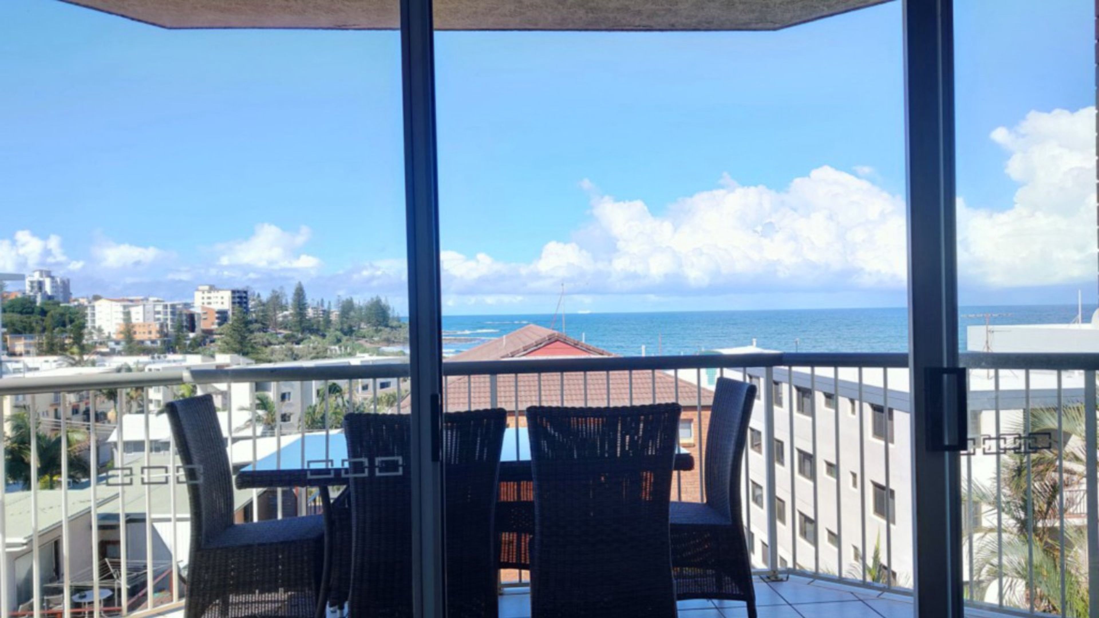 Een balkon met uitzicht op zee, een tafel met stoelen, en gebouwen op de achtergrond.