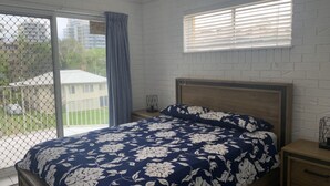 Une chambre à coucher avec un lit, une table de chevet, une fenêtre avec des stores et une vue sur un bâtiment à l’extérieur.