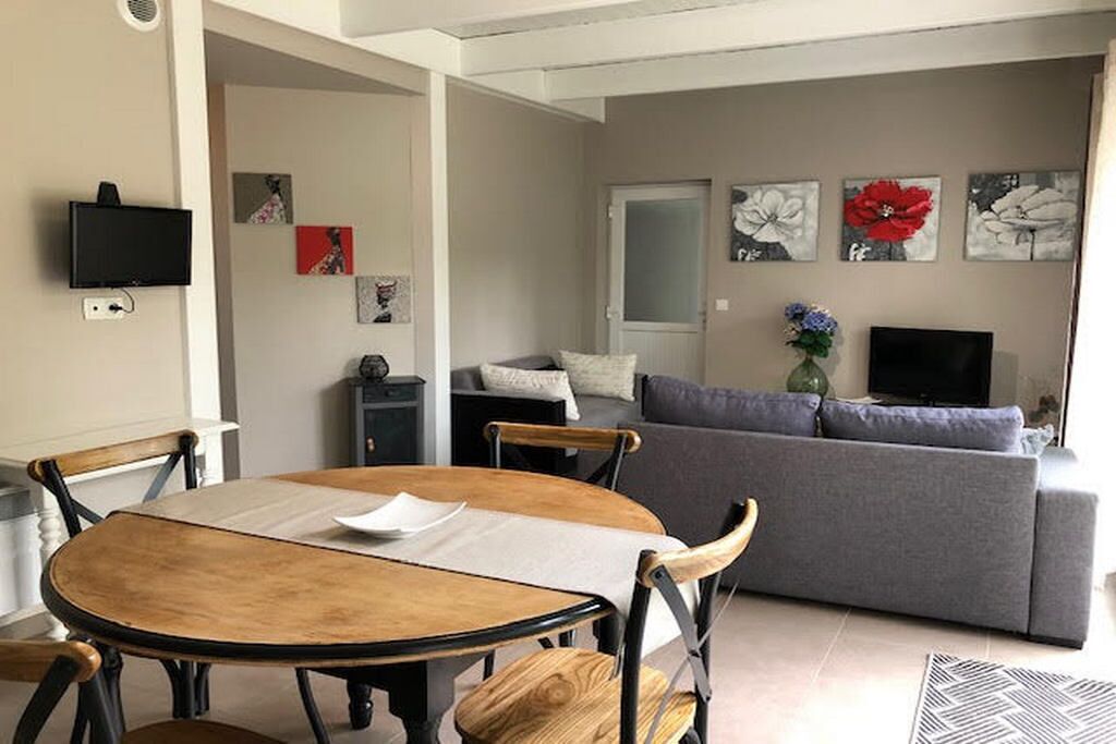 A living room with a grey sofa, a wooden dining table with chairs, a flat-screen TV mounted on the wall, and artwork on the walls.