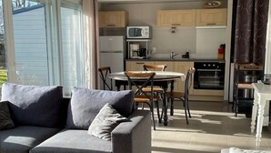 A living room with a grey sofa, a dining table with chairs, and a kitchen area with cabinets and appliances. - Les Cottages du golf 2/3 (LE TRONCHET)