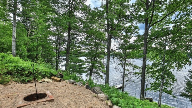 A campsite with a fire pit, surrounded by trees and a lake. - God’s Country Minocqua, Wi. - “Up North” @ Back Bay Cabins on Lake Shishebogama  (Minocqua)