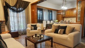 Sala de estar com sofá, poltronas e mesa de centro. Há uma área de jantar com mesa e cadeiras. O ambiente possui armários de madeira e uma cozinha com geladeira.