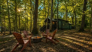 A forest setting with a cabin, a fire pit, and Adirondack chairs.