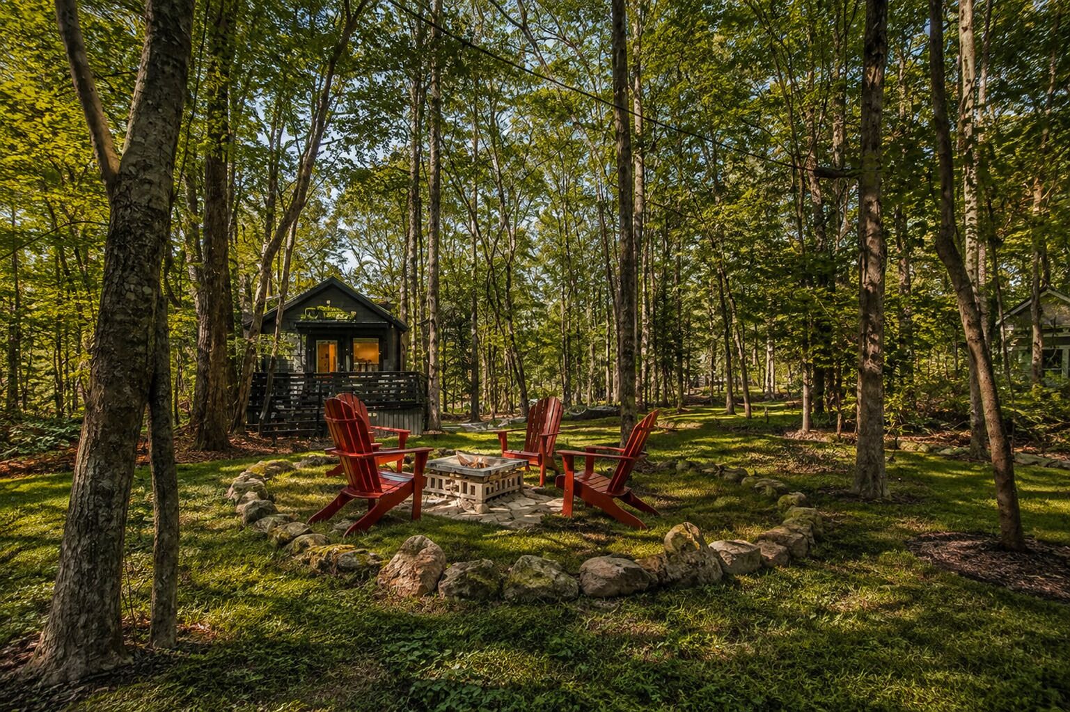 Une cabane entourée d’arbres, avec un foyer et des chaises rouges.