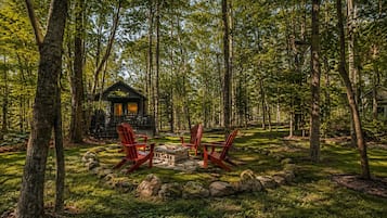 A cabin surrounded by trees with a fire pit and red chairs.