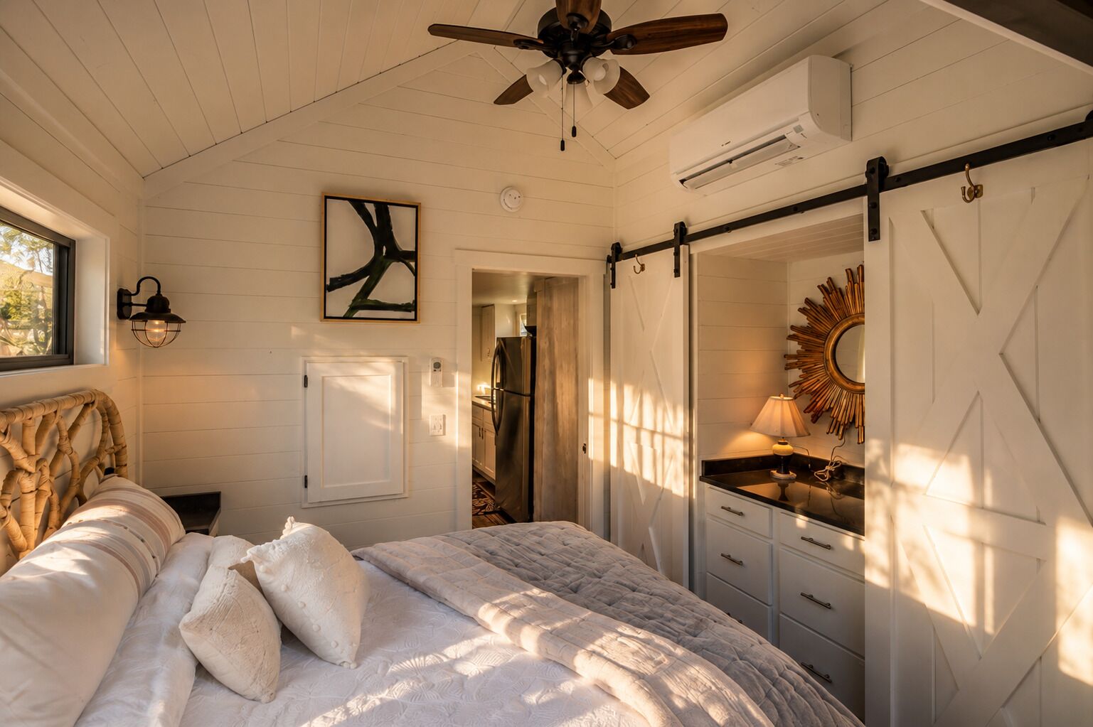 Une chambre à coucher comprenant un lit, un ventilateur de plafond, une lampe fixée au mur, une porte et un miroir orné d’un motif en forme de soleil rayonnant.
