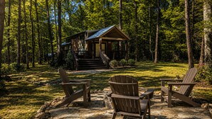 A cabin with a porch and steps leading up to it, surrounded by a forest. There are two Adirondack chairs in the foreground. - The Woods at The Retreat at Deer Lick Falls (Monteagle)