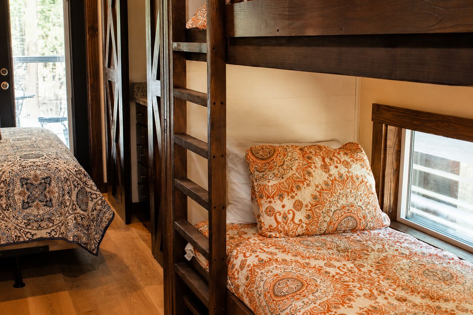 A bunk bed with patterned bedding, a wooden ladder, and a window with a view.