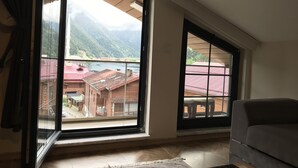A room with large windows offering a view of a mountainous landscape and a building with red roofs. - Anemo Suite (Çaykara)