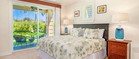 Une chambre avec un grand lit, un ventilateur de plafond, une table de chevet avec une lampe et une vue sur un jardin, accessible par une porte coulissante.
