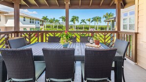 An outdoor patio with a wooden table and wicker chairs, overlooking a tropical landscape with palm trees and a clear sky.