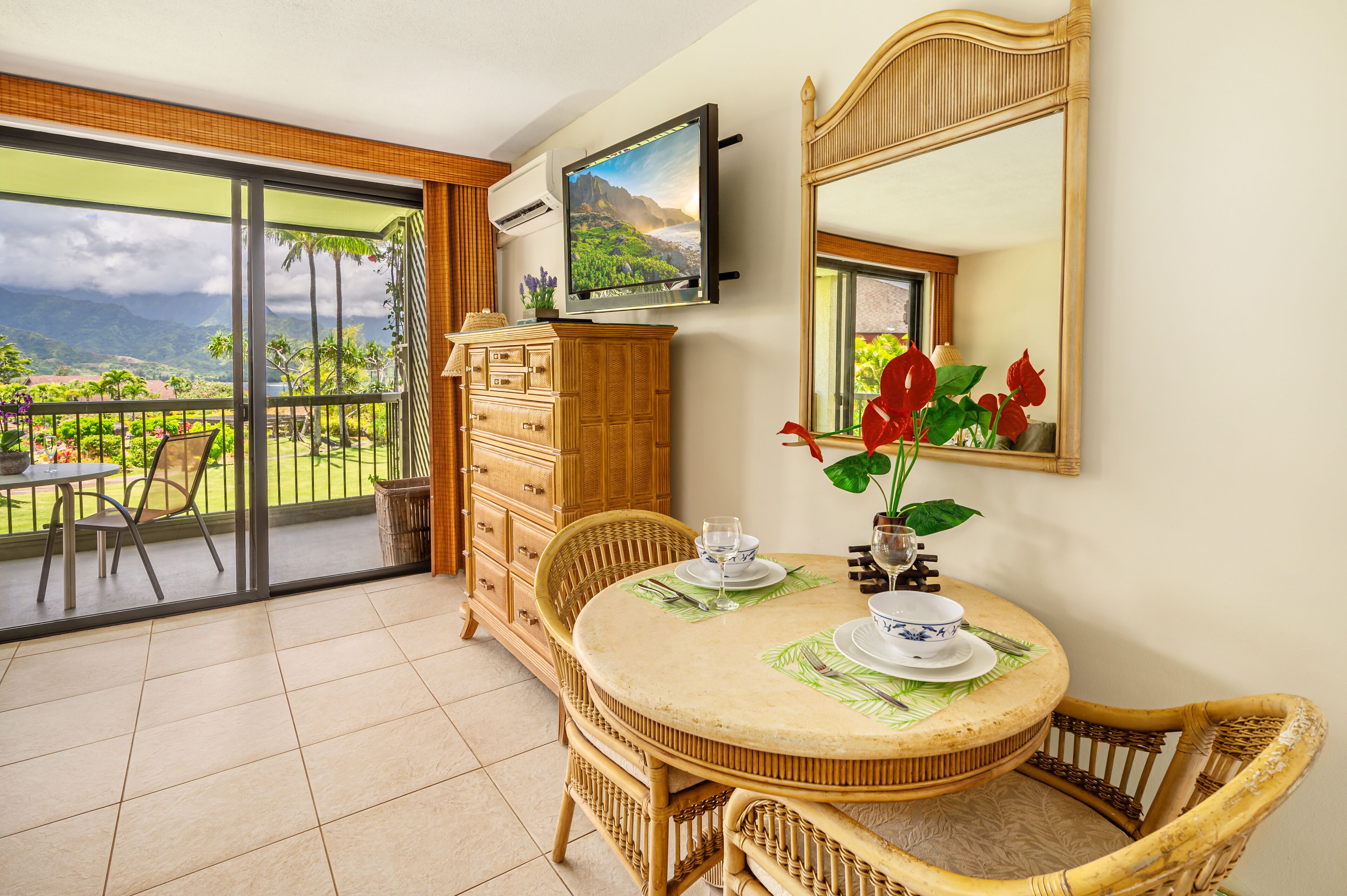 Un coin repas avec une table dressée pour deux personnes, un téléviseur fixé au-dessus de la table et une vue sur un paysage tropical, visible à travers les portes-fenêtres coulissantes.