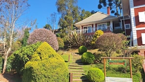 Um jardim bem cuidado, com arbustos aparados e um caminho que leva a uma casa com fachada vermelha e telhado de telhas.