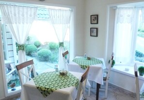 Um quarto com duas mesas redondas, cada uma coberta por uma toalha quadriculada verde e branca. Cadeiras brancas estão dispostas ao redor das mesas. O quarto possui grandes janelas com cortinas brancas e duas quadros emoldurados na parede.