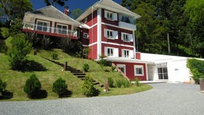 A multi-story building with a red and white facade, a covered driveway, and a landscaped hill with stairs leading to the entrance. - Pousada Mar Deny Campos (Campos do Jordão)