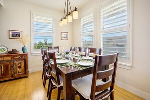 Un coin repas avec une table en bois, dressée pour quatre personnes, entourée de chaises, et offrant une vue sur l’extérieur grâce à de grandes fenêtres équipées de stores.
