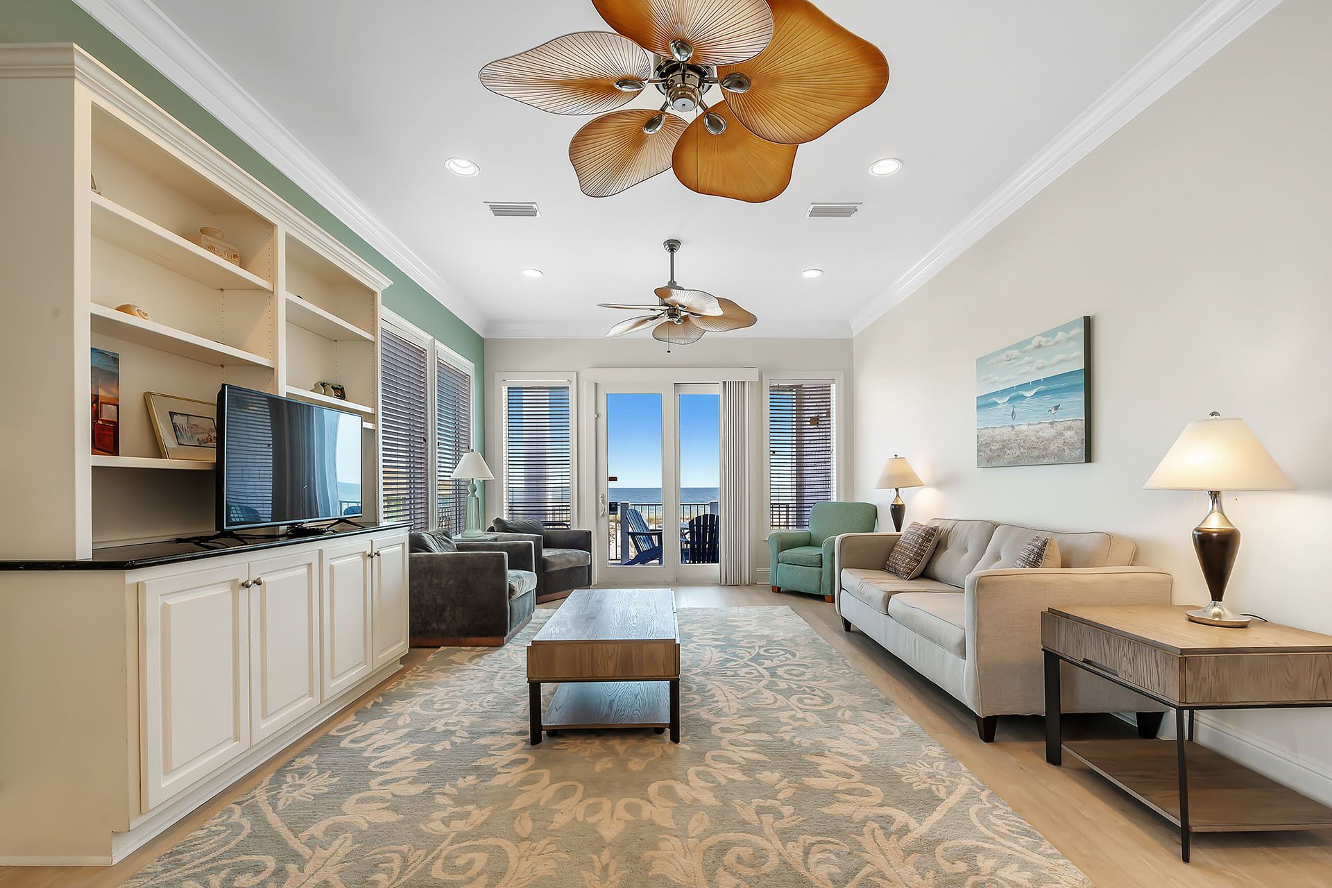 A spacious living area with a large ceiling fan, a television, and a view of the ocean through the sliding glass doors.