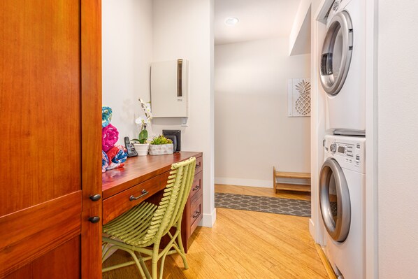 A laundry room with a washing machine, a wooden cabinet, a wicker chair, and a small table with decorative items.