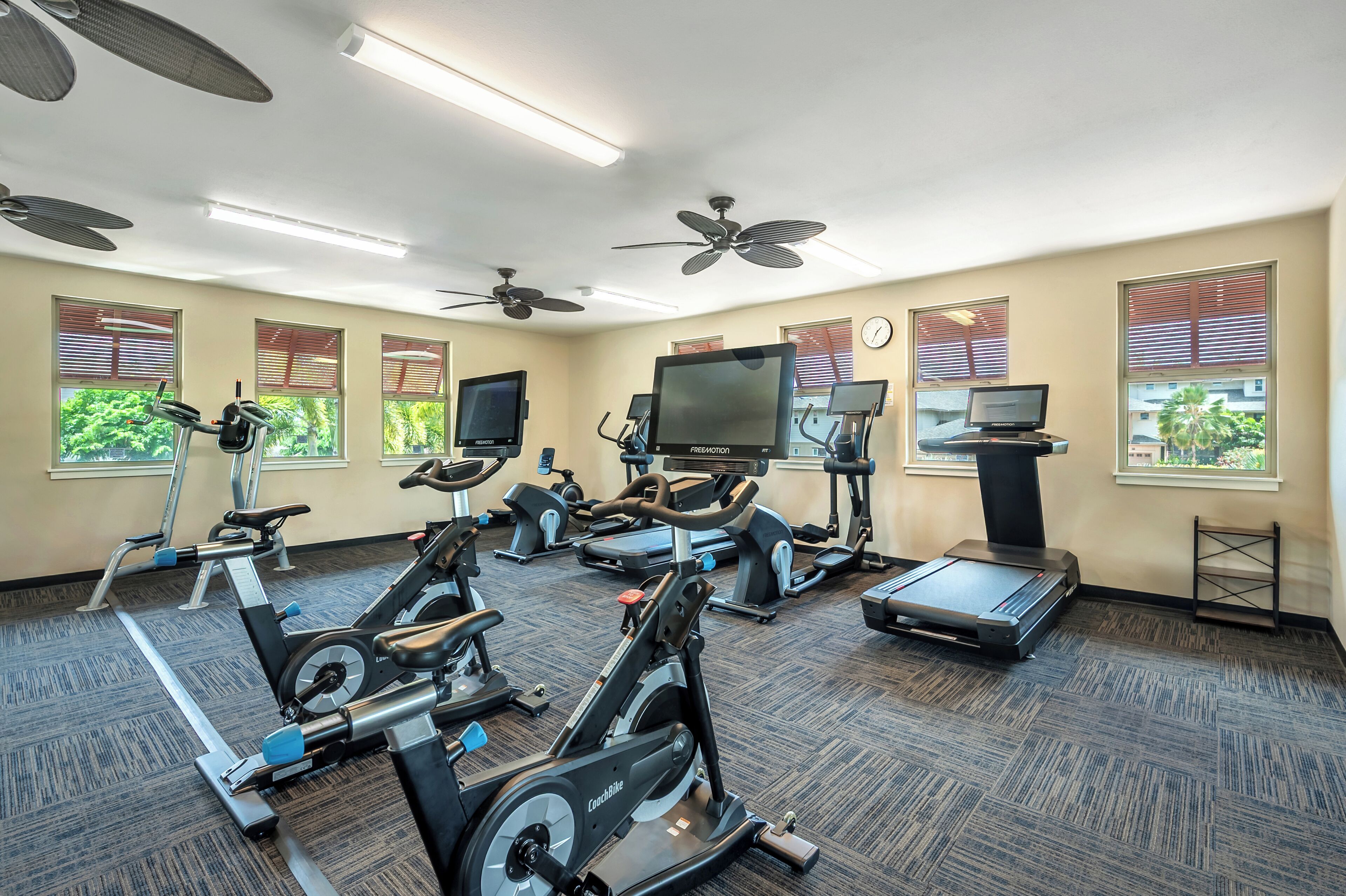 Un gimnasio con varias máquinas de ejercicio, ventiladores de techo y ventanas grandes con persianas.