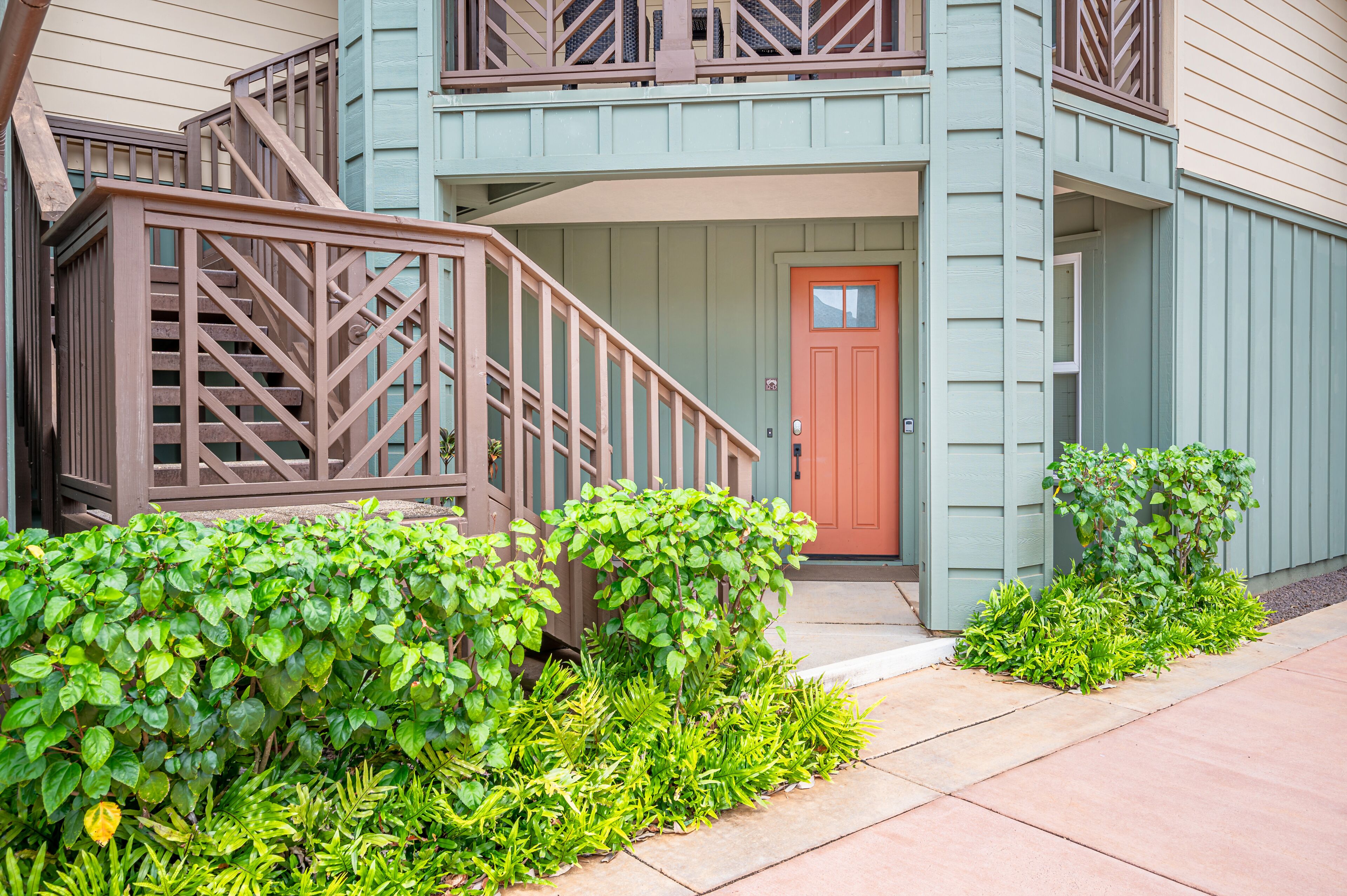 A two-story building with a wooden staircase, a red door, and a small garden area with various plants.