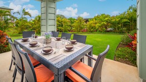 An outdoor dining area with a table set for six, surrounded by wicker chairs with orange cushions, overlooking a well-maintained lawn and tropical plants.