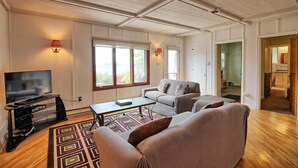 A spacious living room with a large TV, comfortable seating, and a view of the outdoors through the windows.