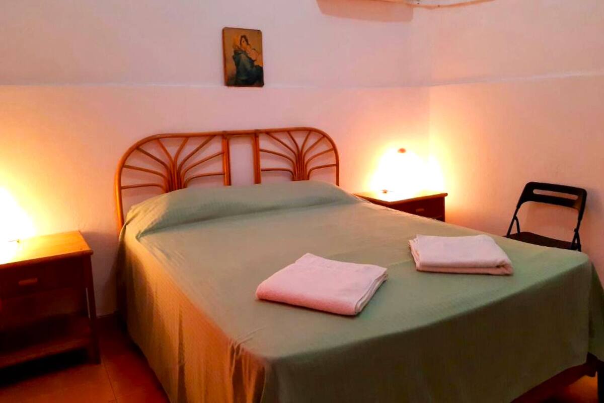 Ein ordentlich bezogenes Bett mit einer grünen Tagesdecke, zwei gefalteten Handtüchern, einem hölzernen Kopfteil, zwei Nachttischlampen und einem kleinen Gemälde an der Wand.