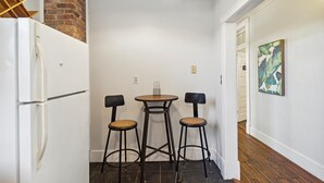 Een compacte keuken met een witte koelkast, een kleine ronde tafel met twee krukken, en een bakstenen muur.