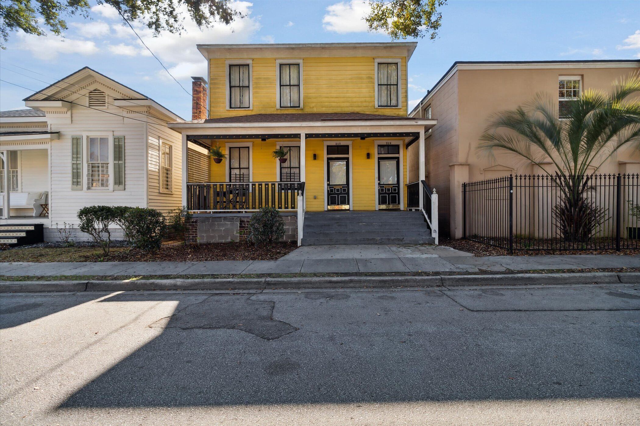 Une maison jaune à deux étages, dotée d’un porche à l’avant, d’un petit jardin et d’une clôture en métal noir.