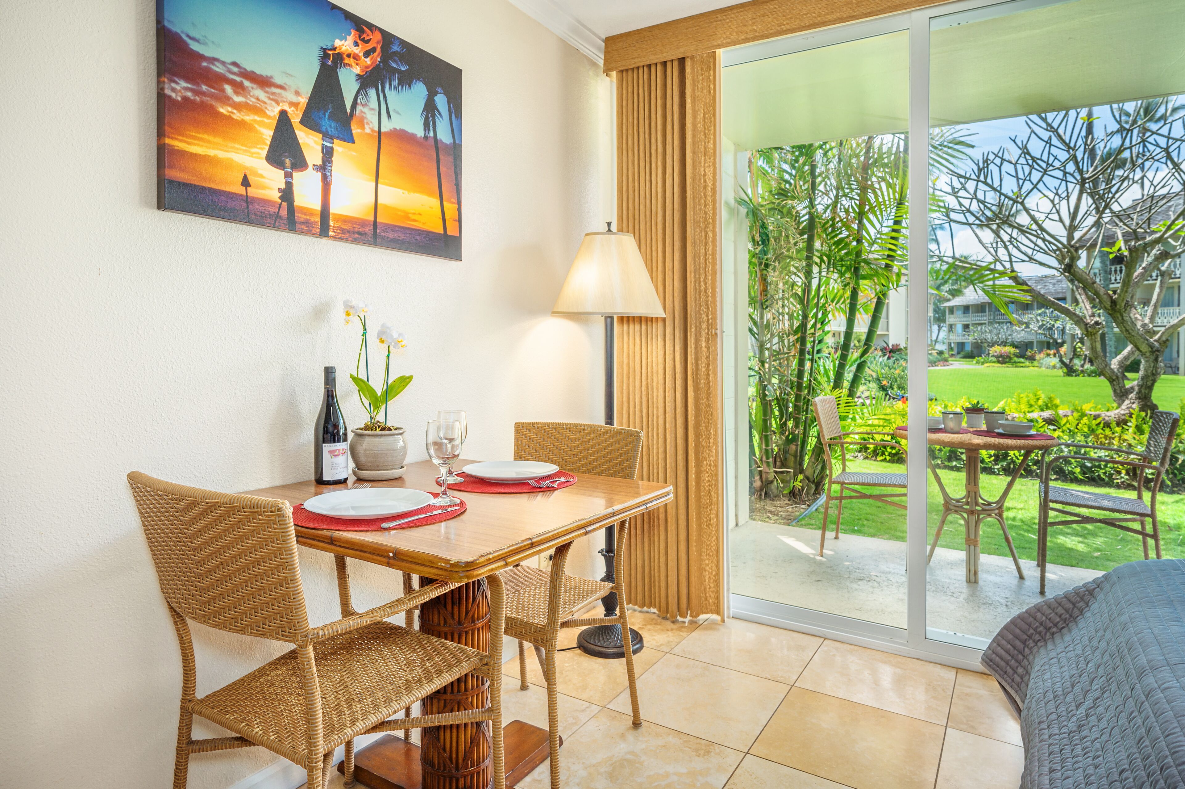 A dining area with a table set for two, a potted plant, and a painting of a sunset.