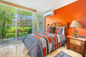 A bedroom with a large sliding glass door to a garden, a bed with a striped quilt, a wooden nightstand, and a lamp. - Islander Family Studio 137, Ground Floor, Steps to Pool and Beach (Kapaa)