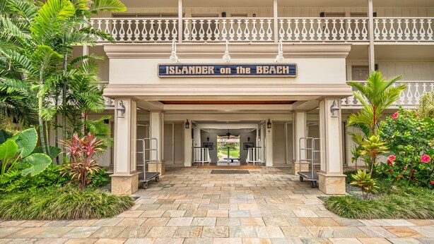 Een hotelentree met een bord waarop staat 'Islander on the Beach', met een overdekte doorgang, stenen vloer en tropische beplanting.