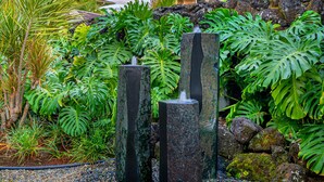 Un jardin tropical avec des palmiers, une fontaine en pierre et un bâtiment en arrière-plan.