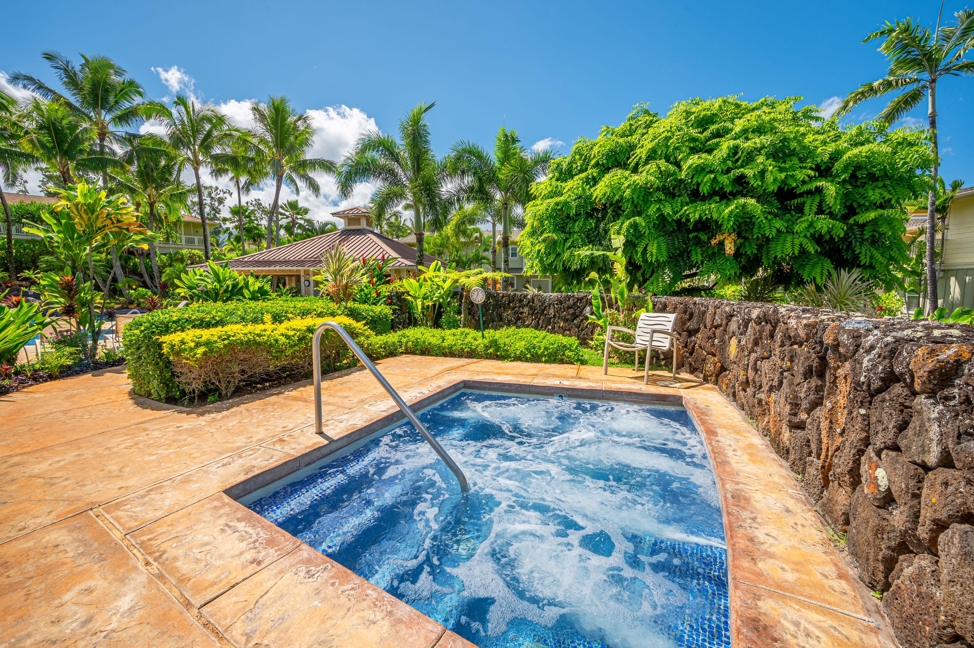 A hot tub surrounded by a stone wall, with palm trees and lush greenery in the background.