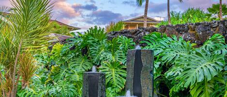 A tropical garden with palm trees, a stone fountain, and a building in the background.