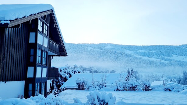 Una cabaña de madera con amplias ventanas y un techo cubierto de nieve, rodeada por un paisaje nevado y colinas boscosas.