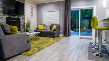 Un salon moderne avec des canapés gris, une table basse et un téléviseur fixé au mur. On y trouve une grande fenêtre avec des stores, un tapis vert et un fauteuil jaune.