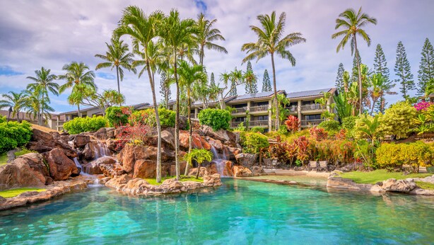 A resort with a clear pool, palm trees, and a waterfall.