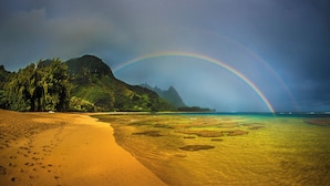 A beach with a rainbow, clear water, and a mountain in the background.