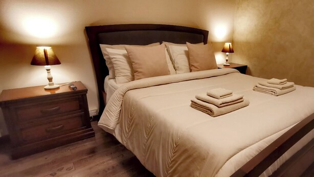 Una cama bien hecha con edredón color beige y almohadas, dos lámparas de noche, una mesita de noche de madera y suelo de madera.