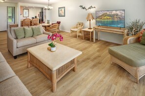 Un salon spacieux avec un canapé, une table basse et des meubles en osier. On y trouve également un coin repas avec des chaises et une table, ainsi qu’un téléviseur fixé au mur.