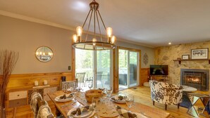 Un comedor con mesa de madera para cuatro personas, una araña y una chimenea al fondo.