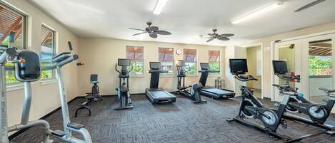 Un gimnasio con diversos equipos de ejercicio, ventiladores de techo y ventanas amplias.