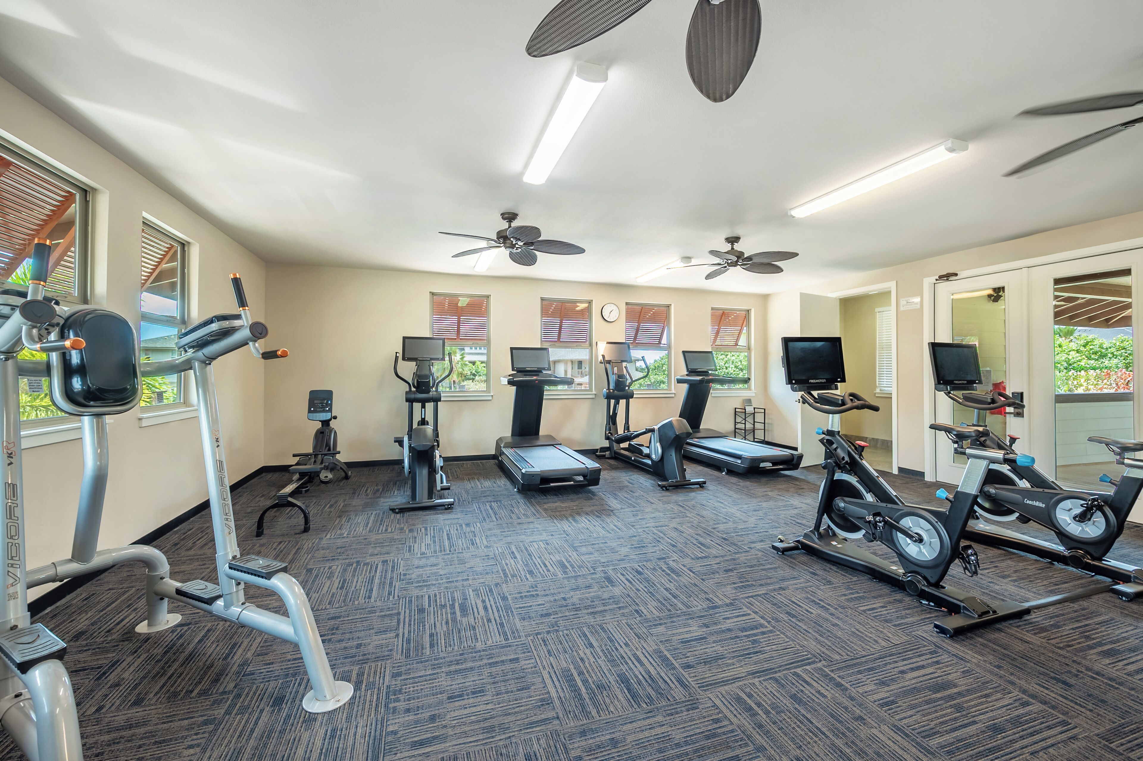 A gym with various exercise equipment, ceiling fans, and large windows.