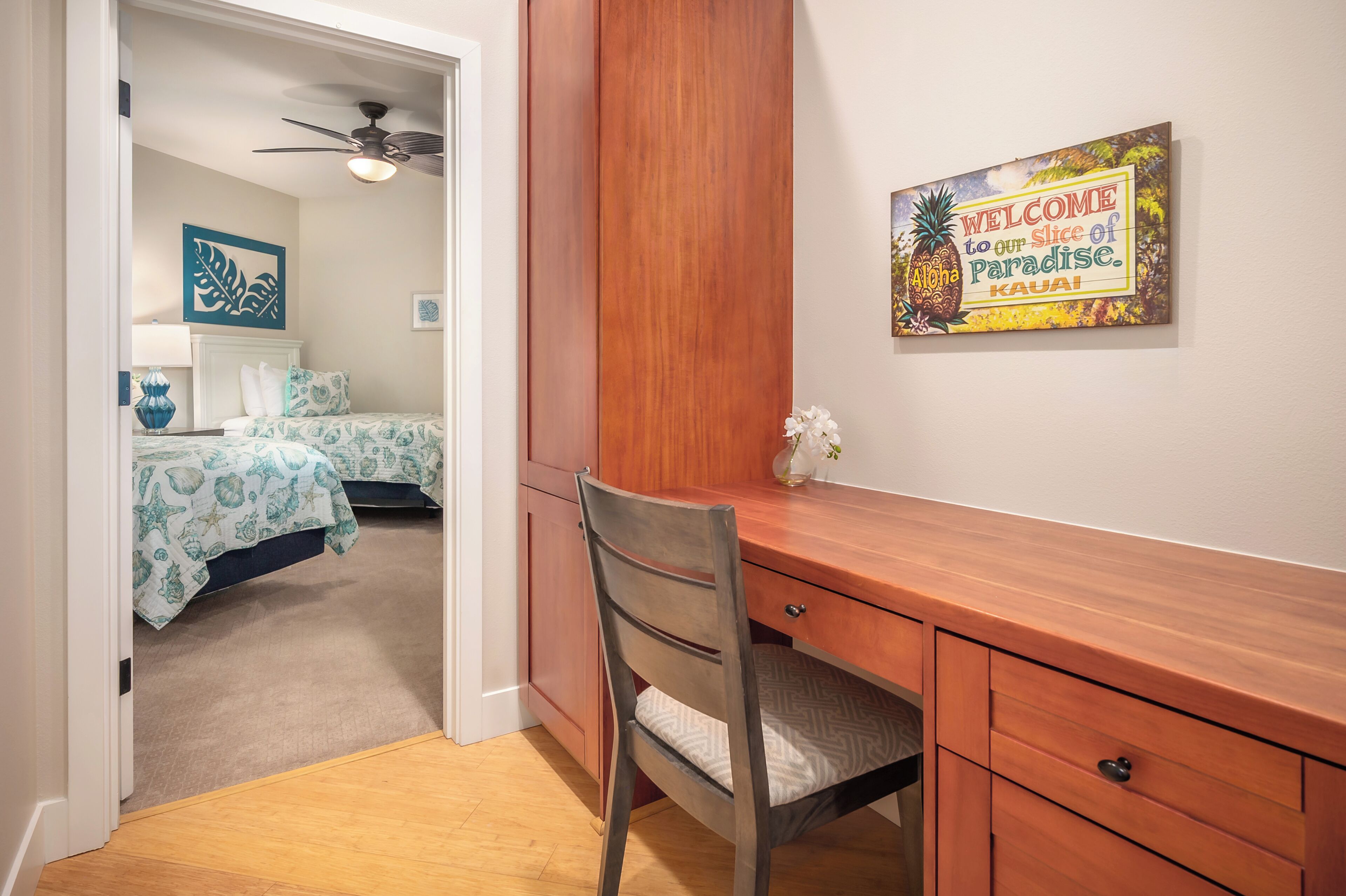 Een slaapkamer met een bed, een bureau en een stoel. Er is een plafondventilator en een bord aan de muur met de tekst: 'Welcome to our slice of paradise. Kauai'.