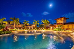 A nighttime resort pool area with a clear view of the pool, palm trees, and a building with a tower.