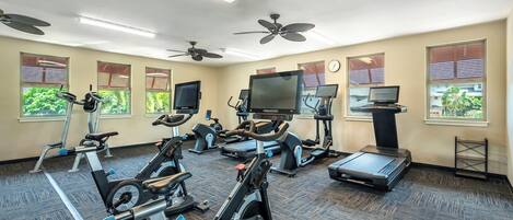Un gimnasio con varias máquinas de ejercicio, ventiladores de techo y ventanas grandes con persianas.