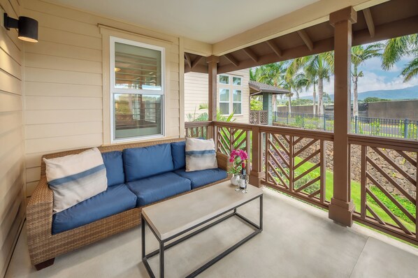 A covered porch with a blue sofa, a coffee table, and a view of a garden with palm trees and a mountain in the background.
