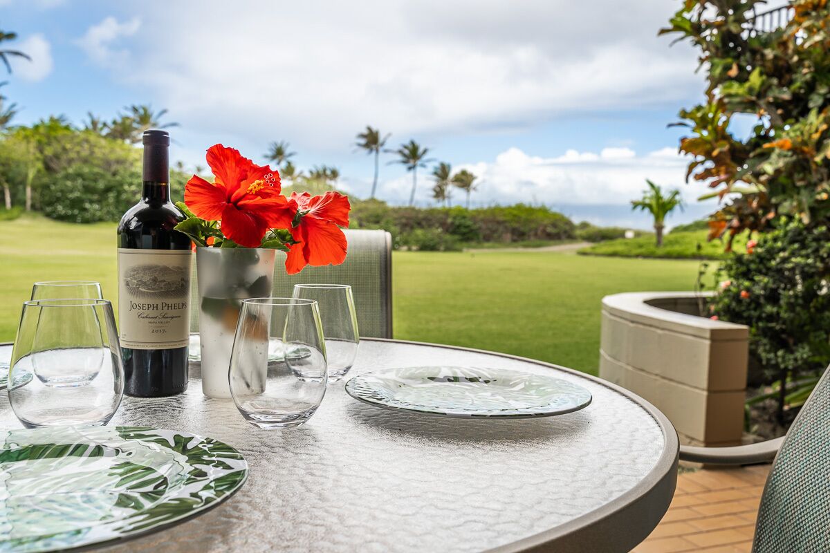 Mesa montada ao ar livre com uma garrafa de vinho, copos e um vaso com uma flor vermelha.