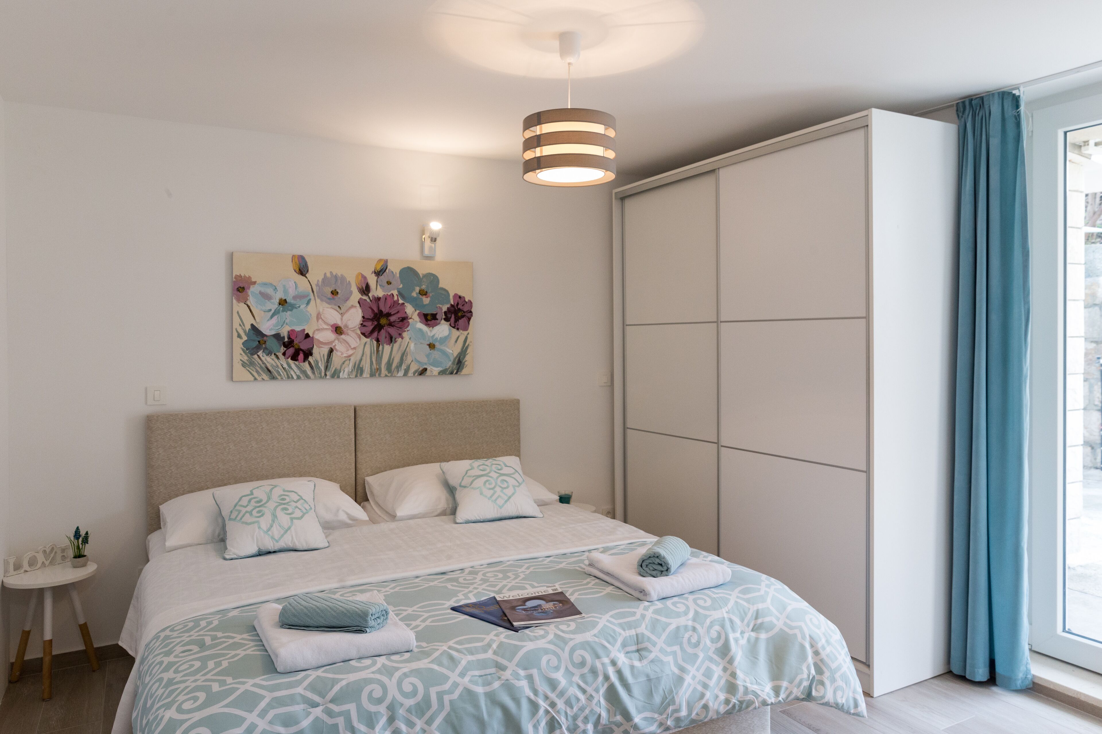 A bedroom with a bed, a nightstand, a painting, a wardrobe, and a window with curtains.